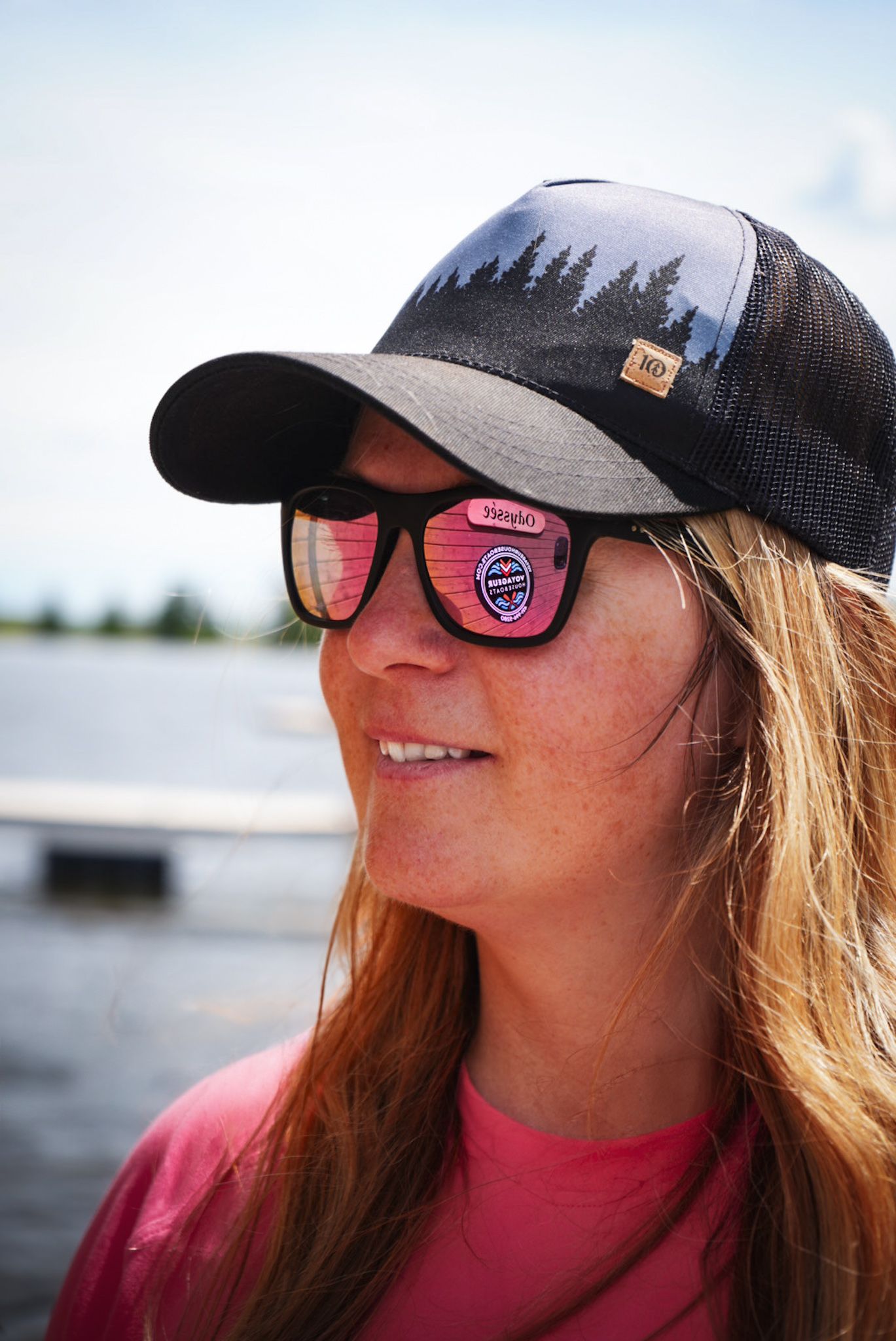 Outdoor enthusiast wearing trucker hat with forest skyline design and pink reflective sunglasses.