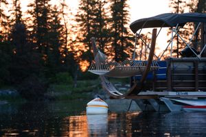 Hammock silhouetted against sunset with pine trees and boats on tranquil lake.