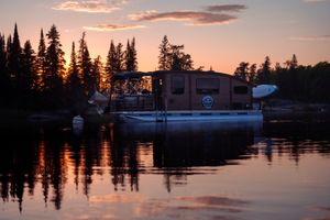 Houseboat silhouetted against sunset with pine trees reflecting on calm lake waters.