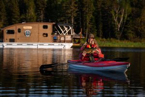 Red kayak floating near houseboat on serene lake surrounded by pine forest.