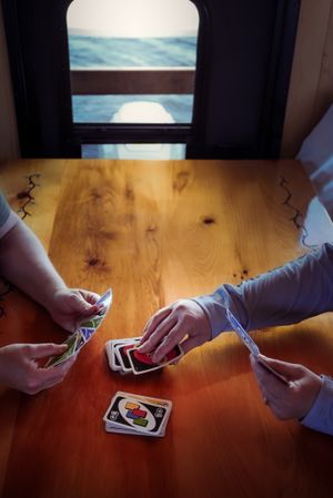 People playing card game Uno at wooden table with ocean view through window.