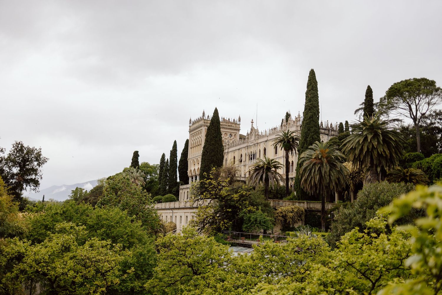 Ancient Mediterranean villa with cypress trees and palm gardens sits atop a hill under cloudy skies in romantic setting.