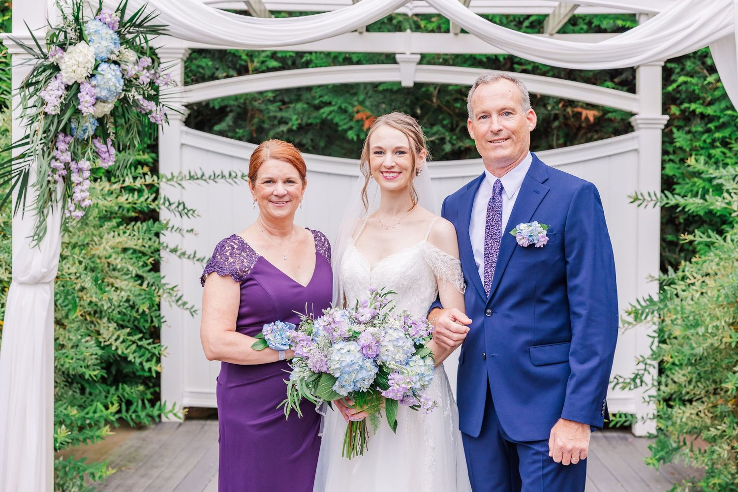 Three people in formal attire stand beneath a decorated garden arch, gathering for a wedding photo moment.