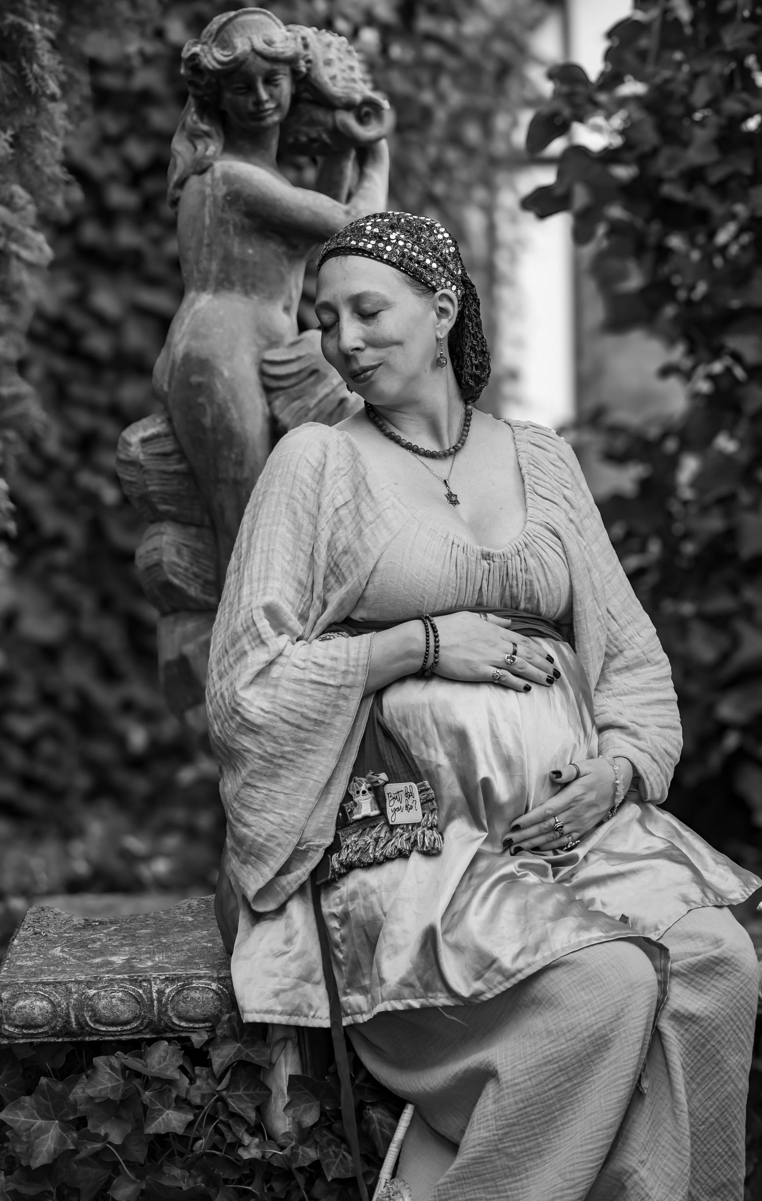 Black and white photo of a pregnant woman in medieval dress sitting near a stone statue in a garden.
