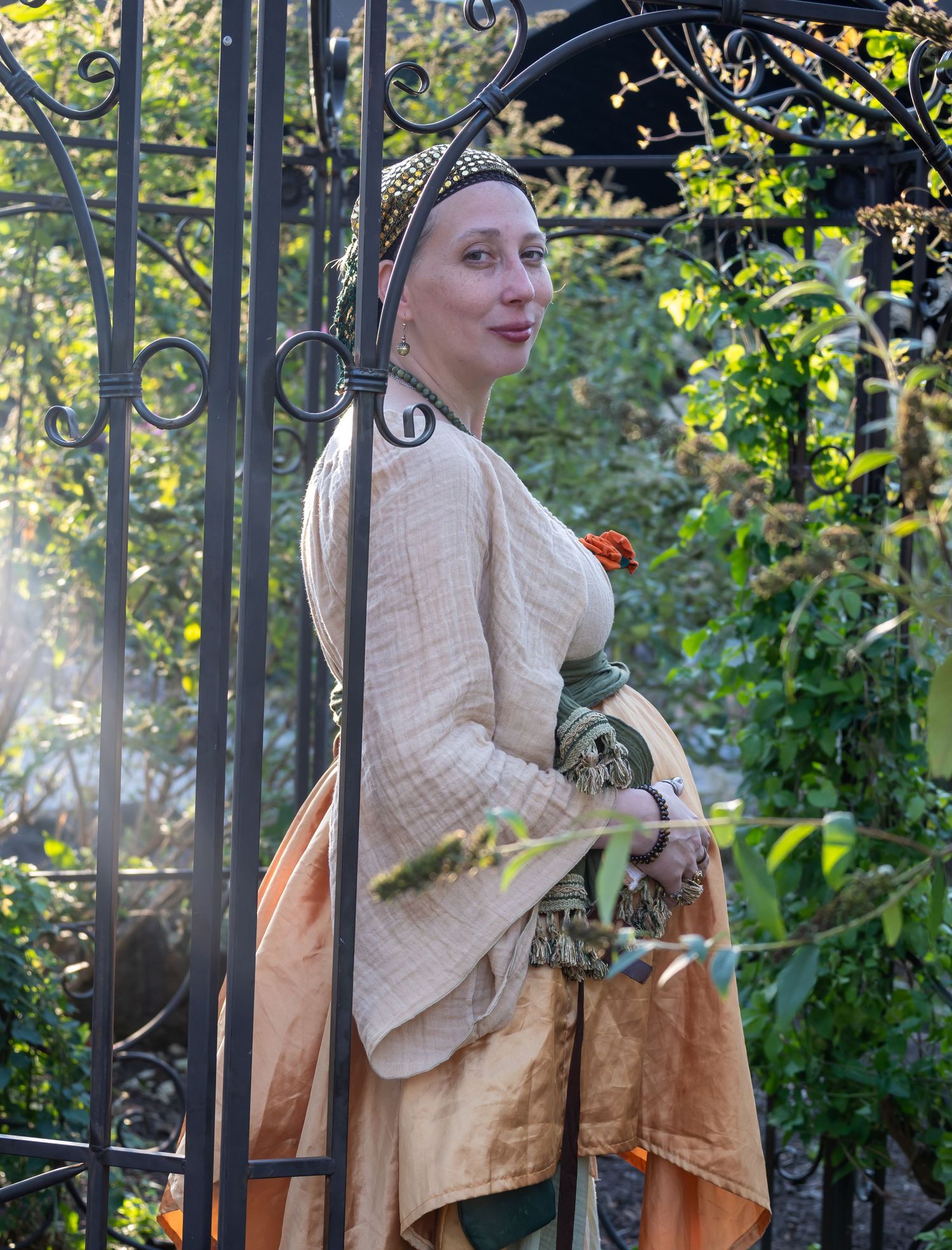 Woman in medieval Renaissance costume stands by ornate iron gate in sunlit garden wearing cream and green gown.
