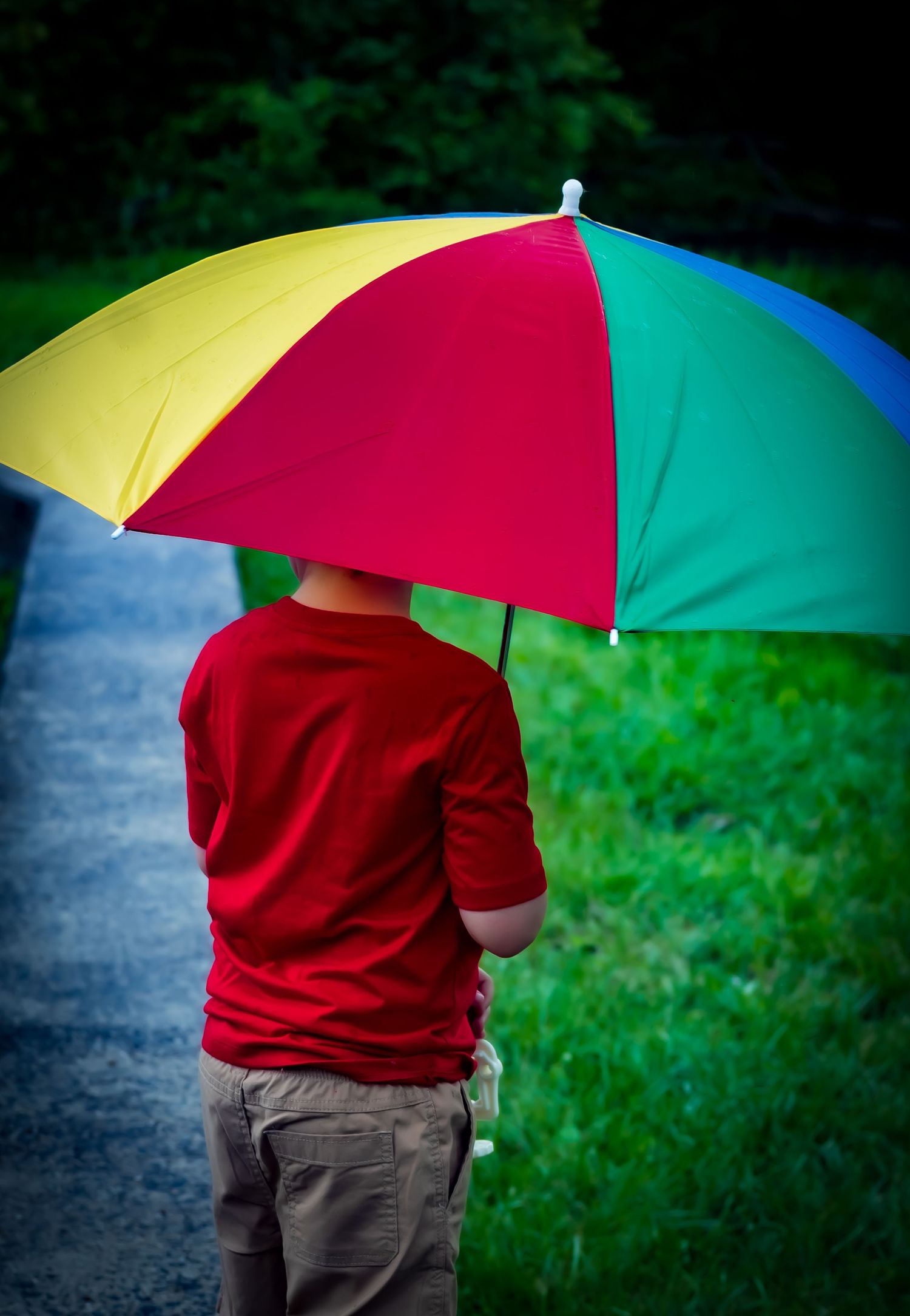 Person in red sweater and brown pants holds colorful rainbow umbrella against blurred natural background in rainy setting.