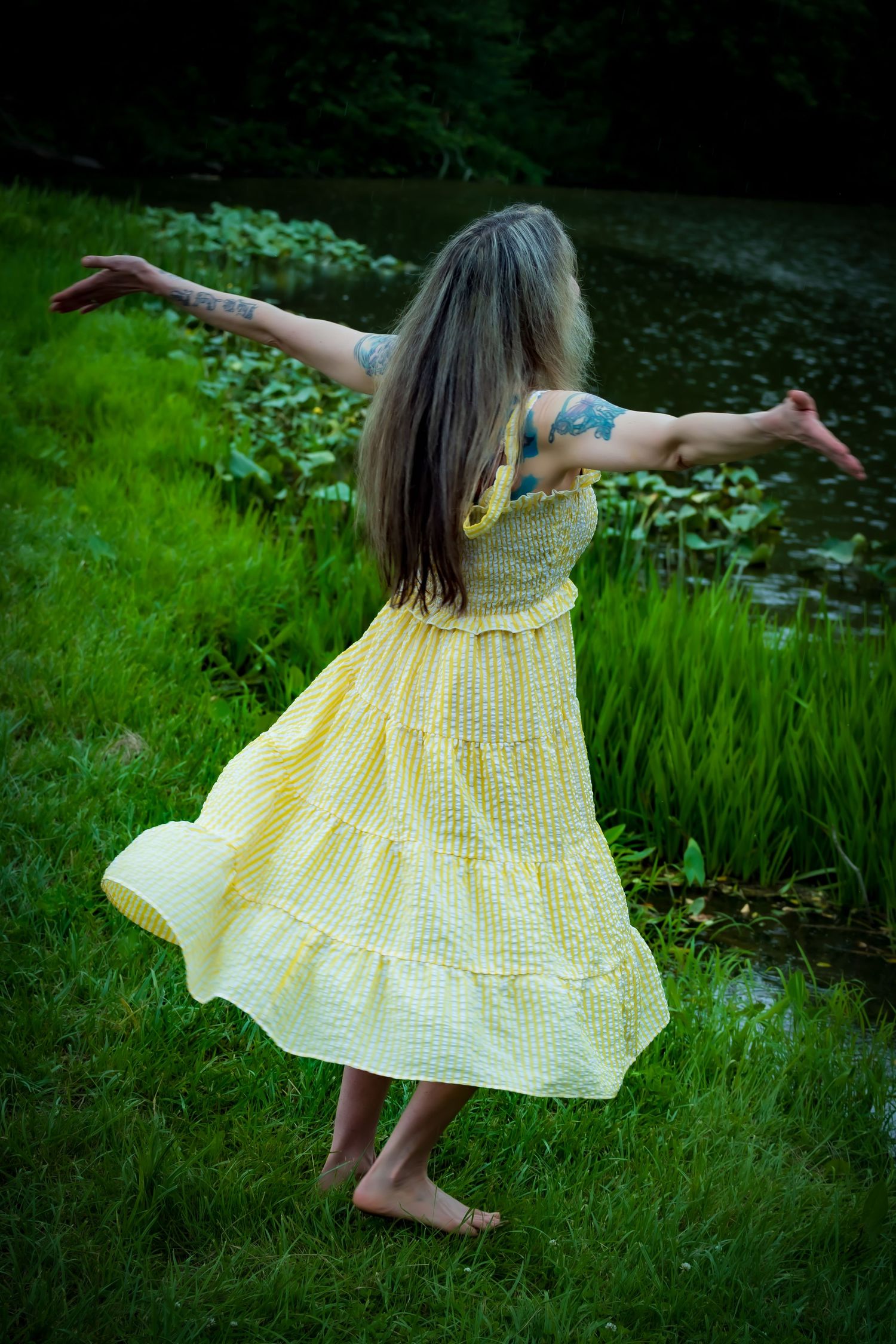 A long-haired dancer in a flowing yellow sundress twirls barefoot on green grass near a pond.