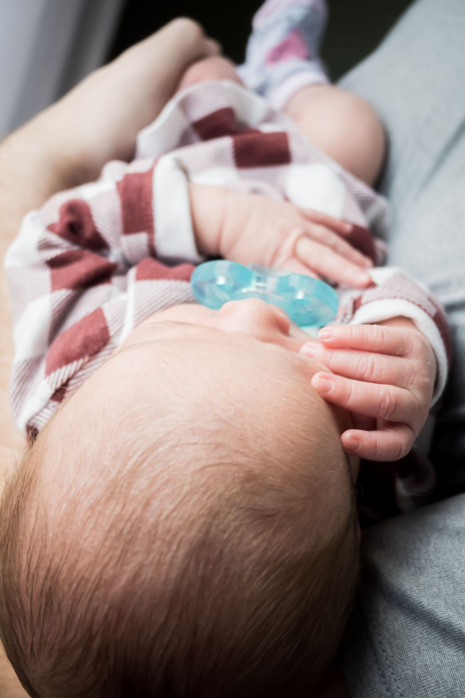 Newborn baby in striped onesie holding a blue pacifier while lying on a caregiver's lap.