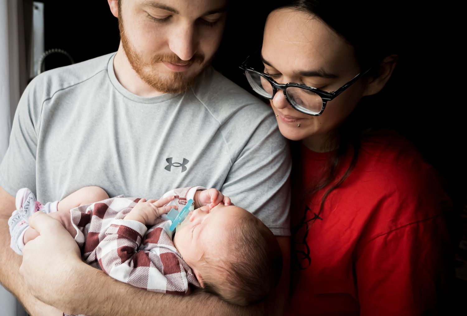 New parents lovingly gaze at their newborn baby, mother in red top and father in gray shirt holding infant.