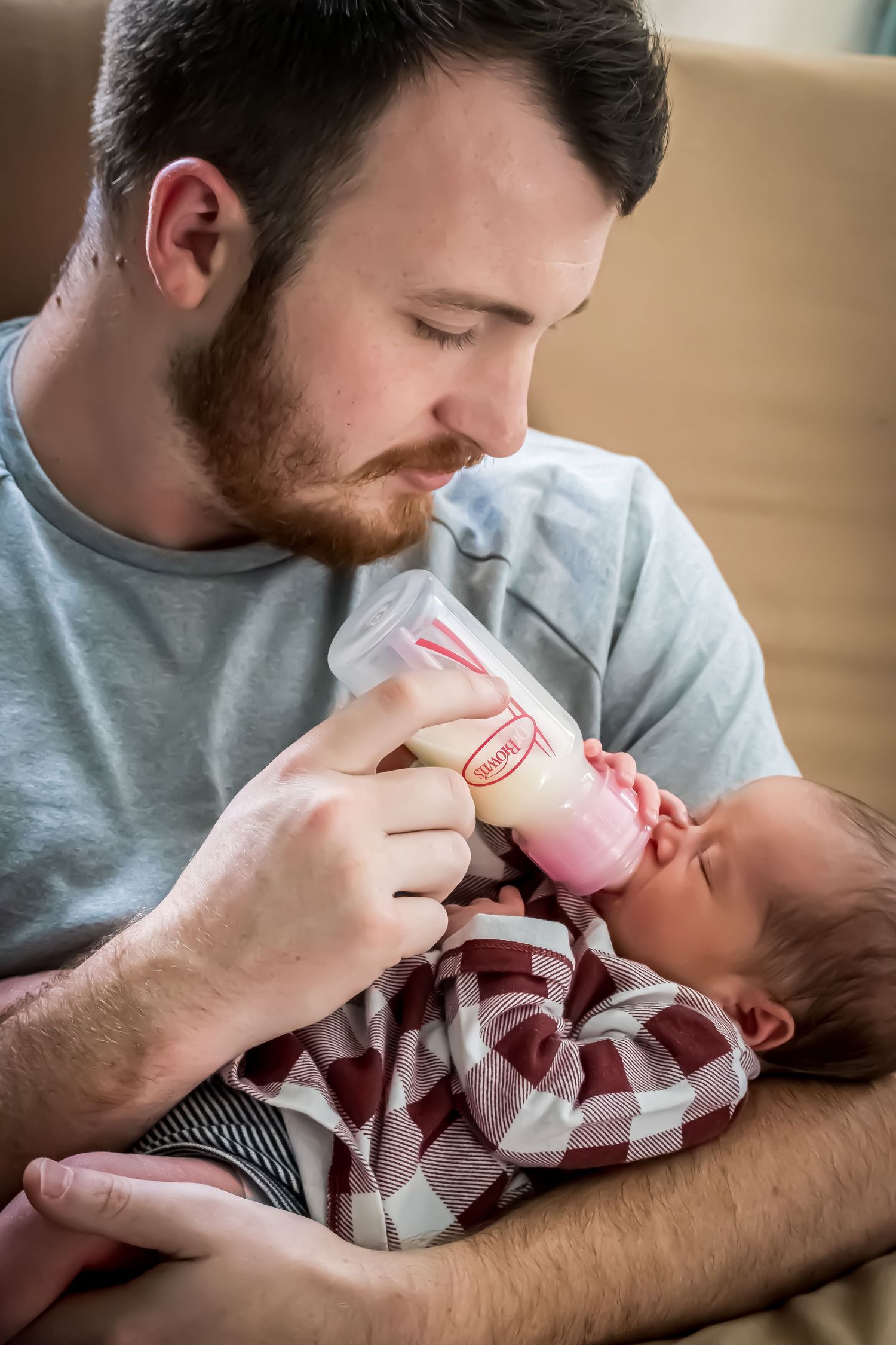 A young bearded man tenderly bottle-feeds a newborn baby dressed in a plaid outfit while cradling it in his arms.