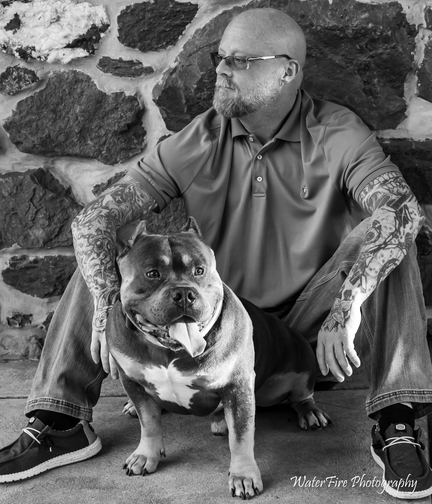 Tattooed bald man with glasses crouches beside a bulldog against a stone wall in a black and white portrait photo.