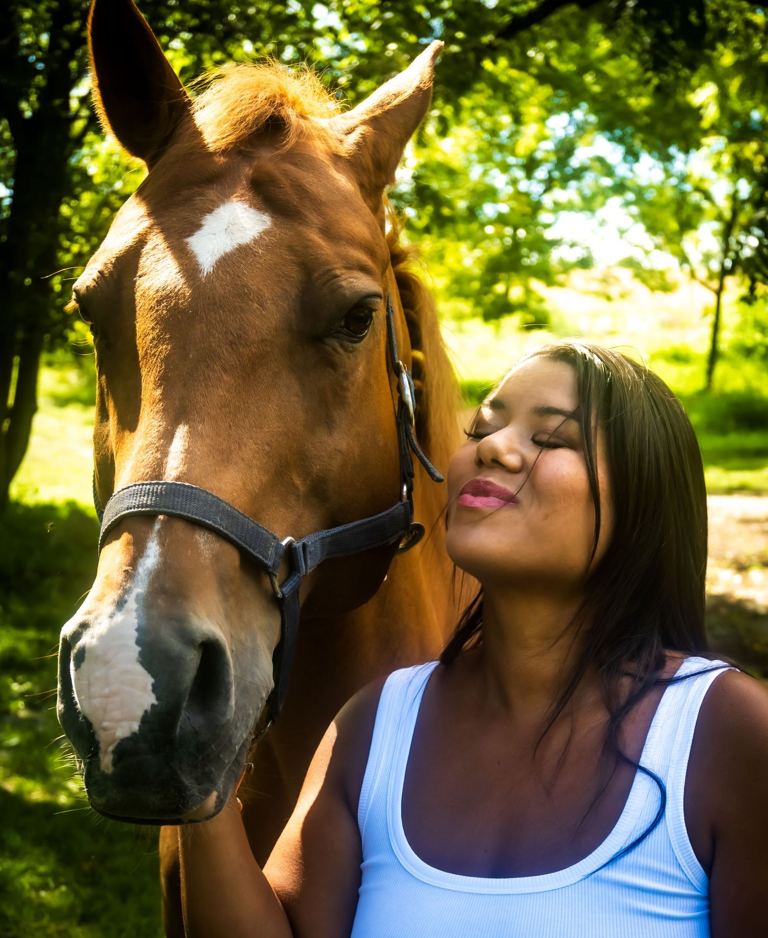 A young woman in a white tank top puckers up to kiss a chestnut horse with a blaze marking outdoors.
