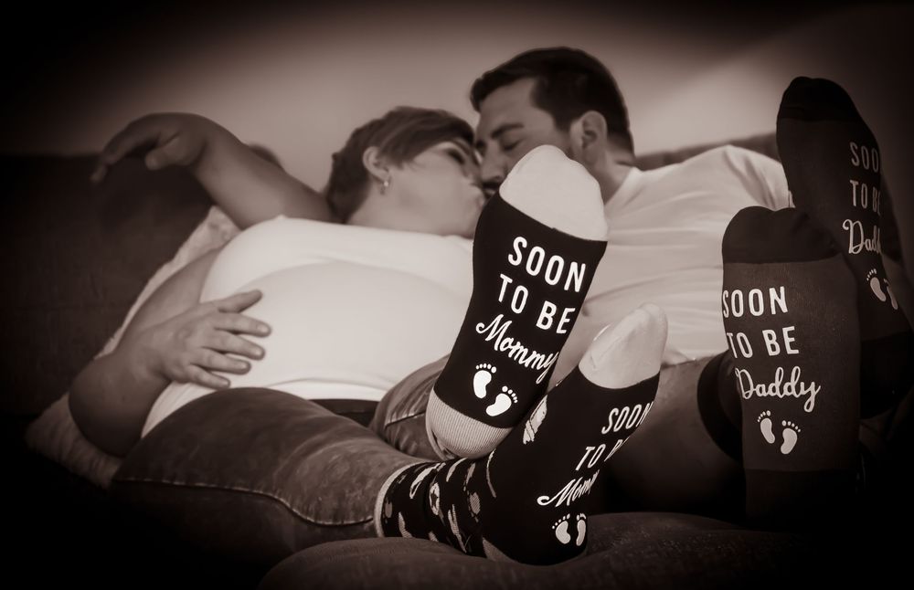 A couple lying down wearing matching 'Soon to be Mommy' and 'Soon to be Daddy' black socks in vintage sepia tones.