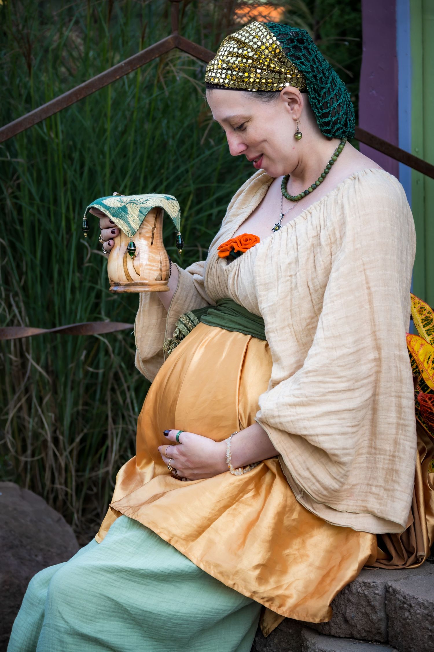 Pregnant woman in medieval Renaissance costume holding decorative dragon cup while sitting outdoors.