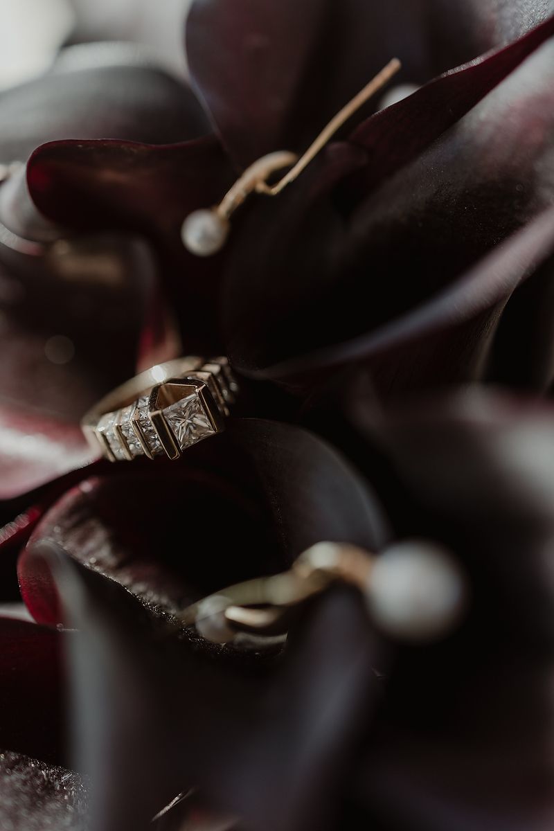 Diamond ring and pearl drop earrings nestled among dark burgundy calla lily petals in a moody close-up shot.