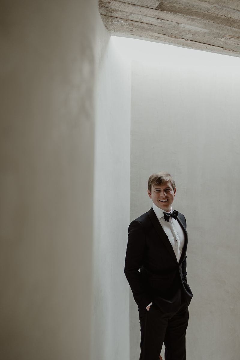 Smiling groom in black tuxedo and bow tie stands with hands in pockets against white wall.