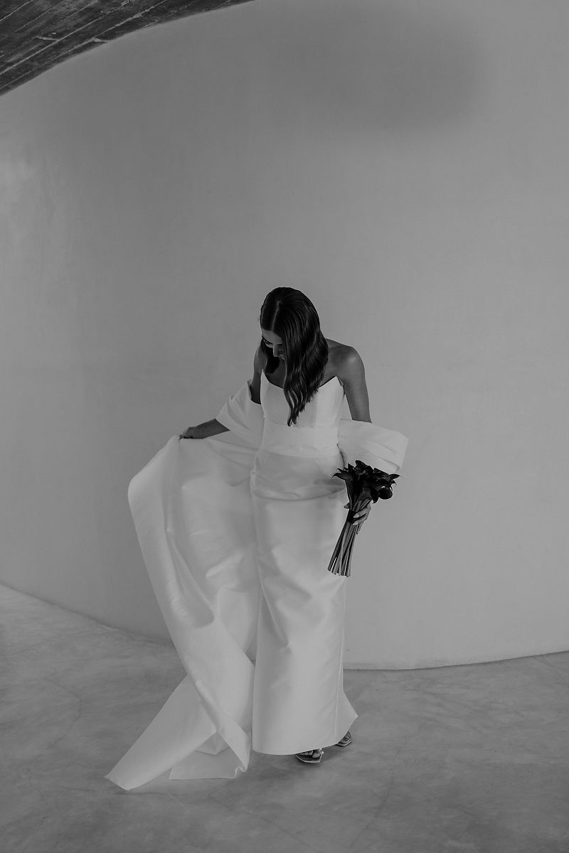 Black and white photo of a bride in a flowing strapless gown holding dark florals in a minimalist white studio space.