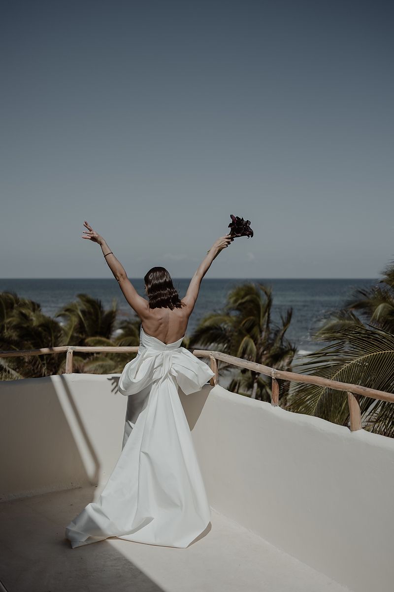 Bride in white backless gown with bow raises arms joyfully on terrace overlooking tropical beach and palm trees.