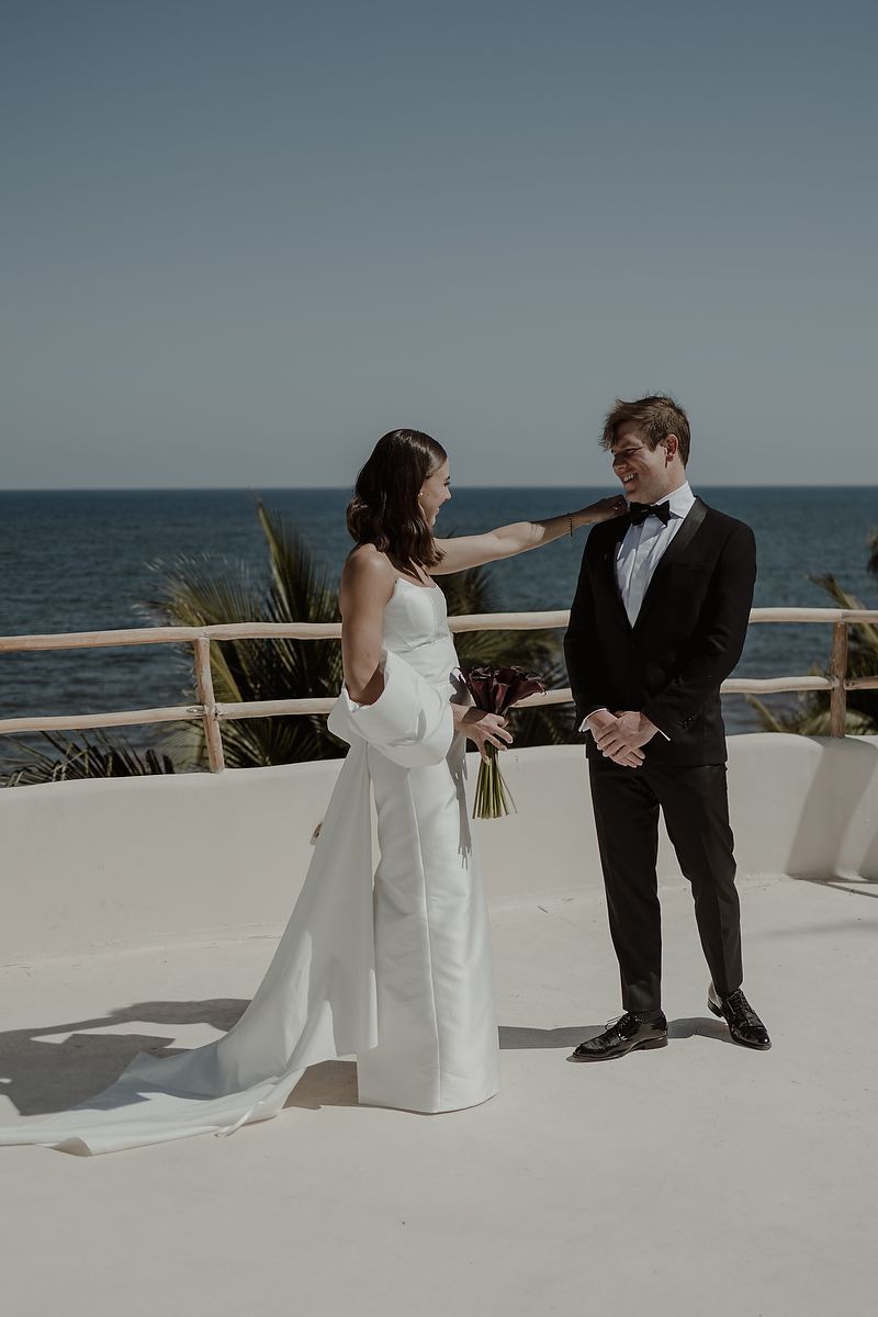 Bride in white gown touches groom's face during oceanfront wedding ceremony on white terrace.