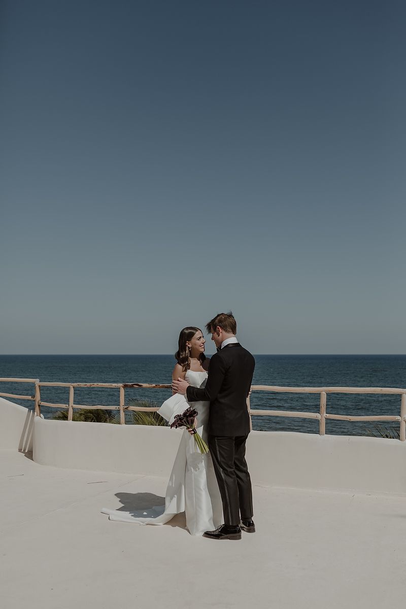 Bride and groom embrace on white terrace overlooking the ocean under a clear blue sky on their wedding day.