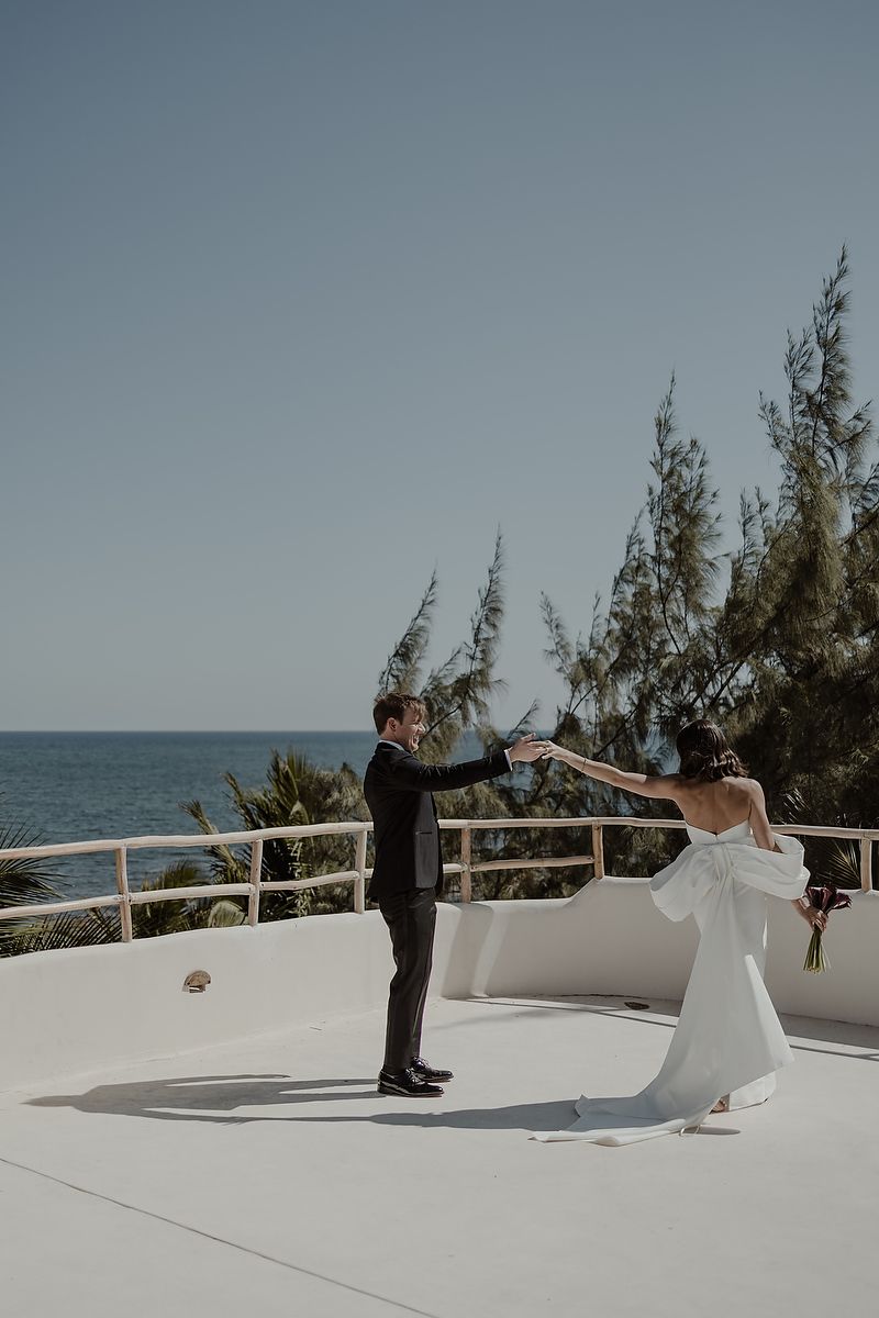 Groom spins bride in white dress on rooftop terrace with ocean and trees in background.