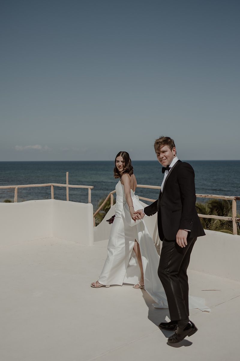 Elegant bride and groom holding hands on a white rooftop terrace with ocean views on their wedding day.