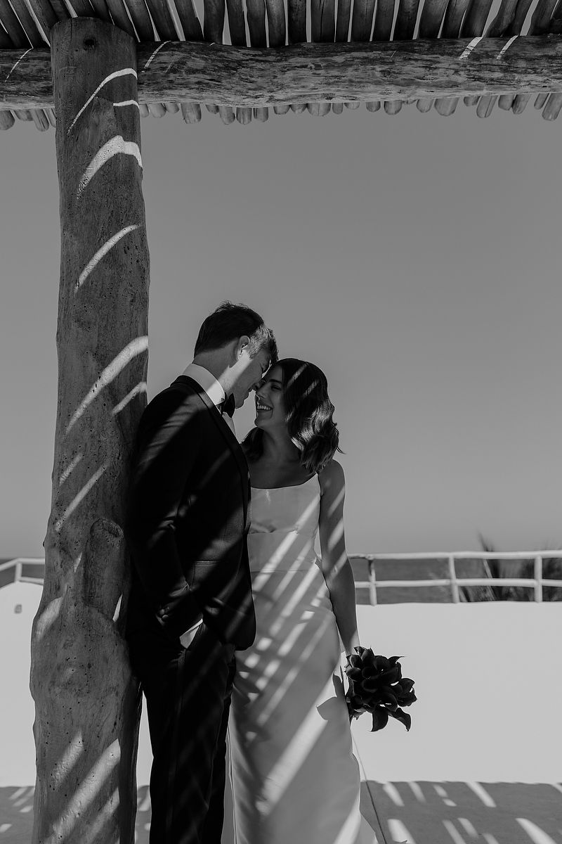 Black and white photo of newlyweds sharing a kiss under a rustic wooden pergola, bride holding a dark floral bouquet.