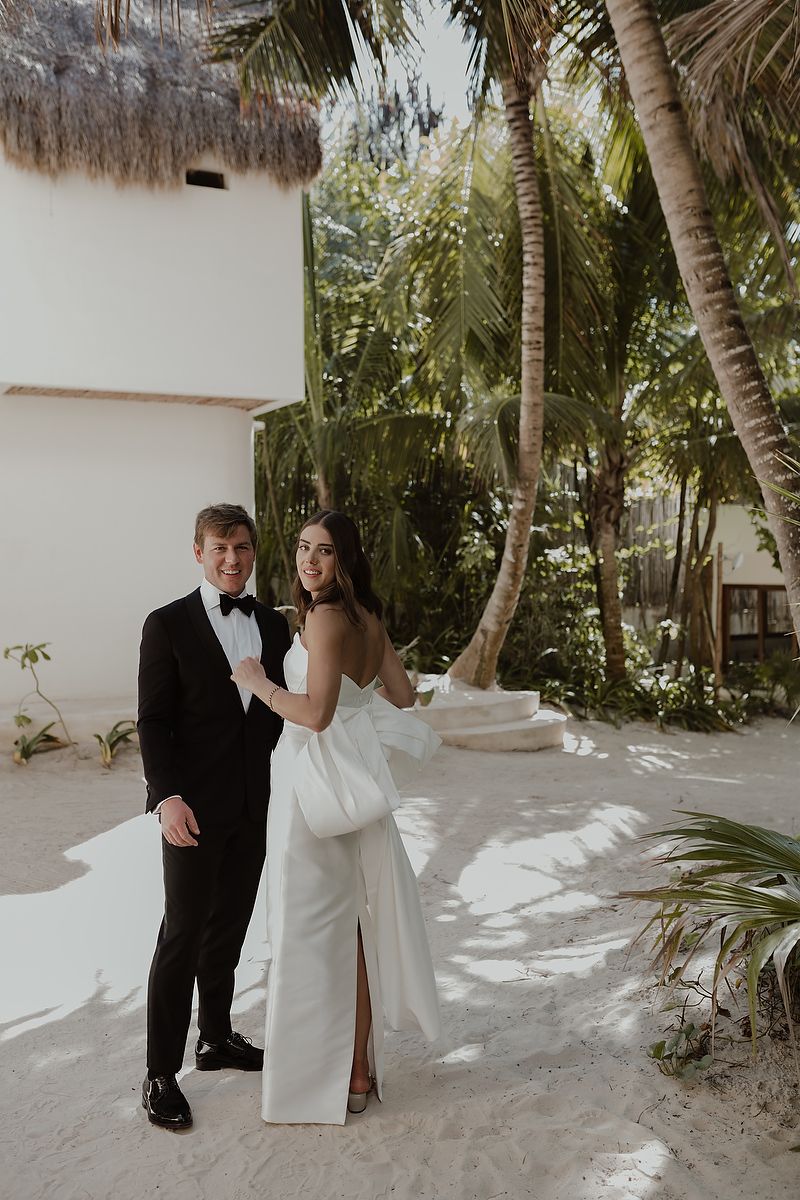 Bride and groom posing together on sandy tropical grounds with palm trees, bride in white gown, groom in black tuxedo.