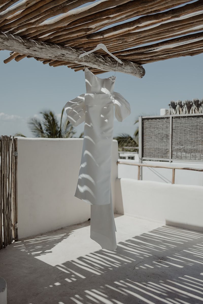 White dress hanging on a hanger under a rustic wooden pergola on a sunny rooftop terrace.