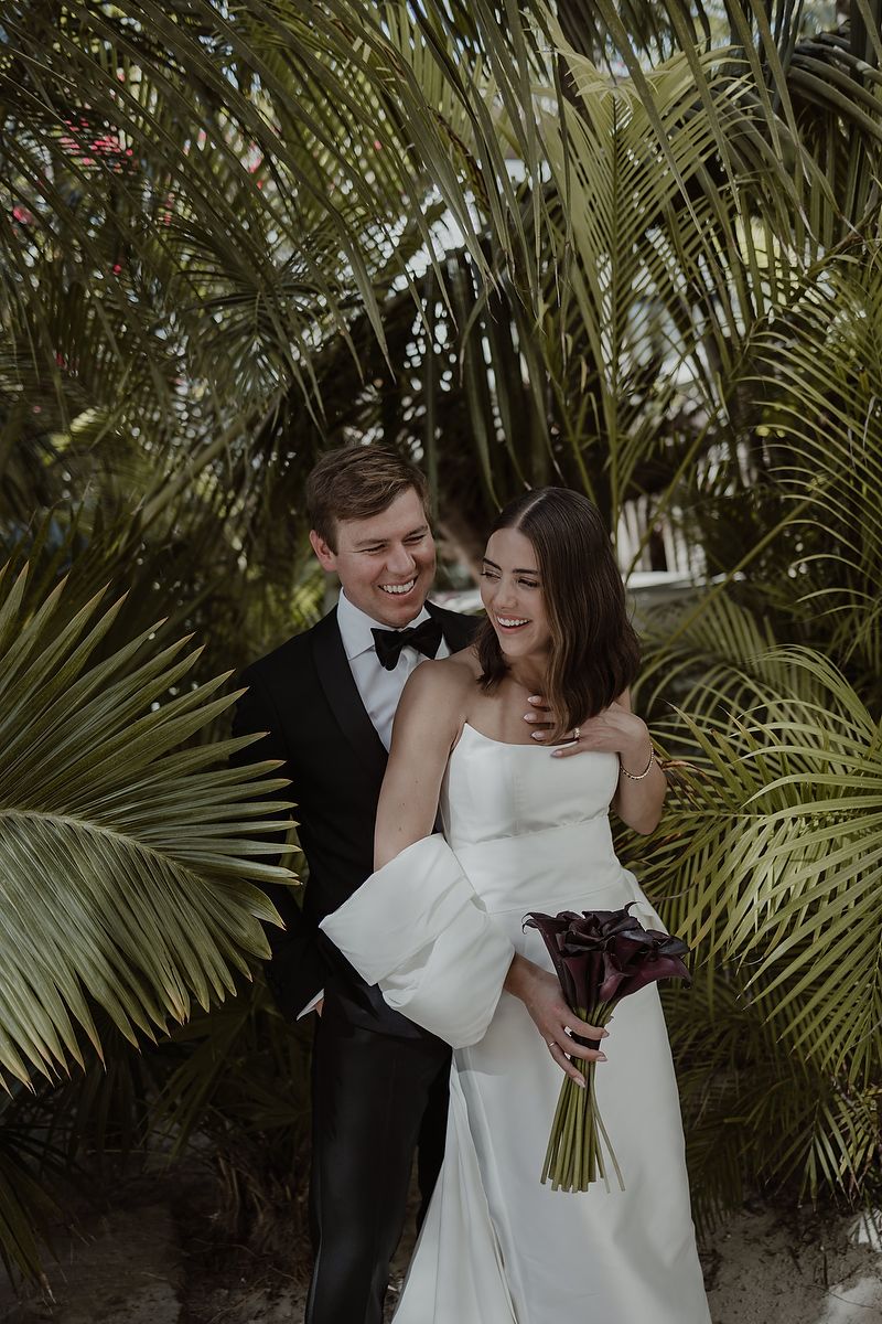 Joyful groom in black tuxedo embracing bride in strapless gown with palm trees in background.