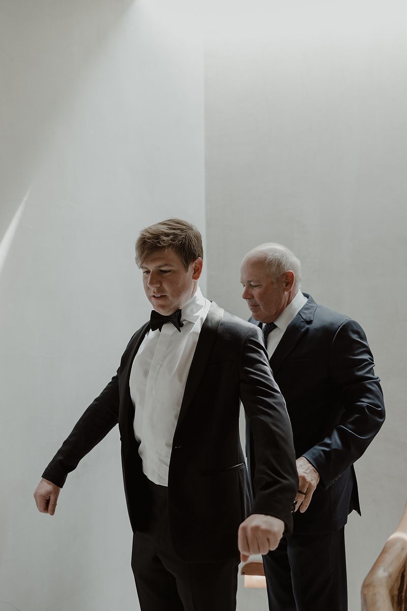 Groom in black tuxedo and bow tie getting ready with older man in dark suit in bright white room.