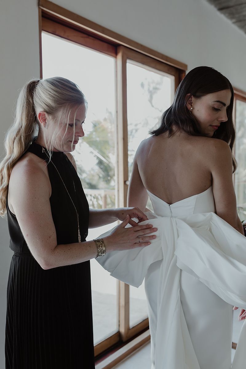 A bridesmaid in black helps a bride in a strapless white gown with her dress near large windows.
