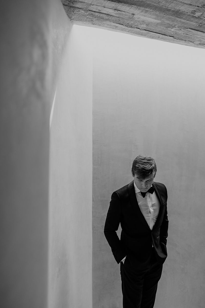 Elegant groom in a black tuxedo with bow tie stands contemplatively in a minimalist architectural space.