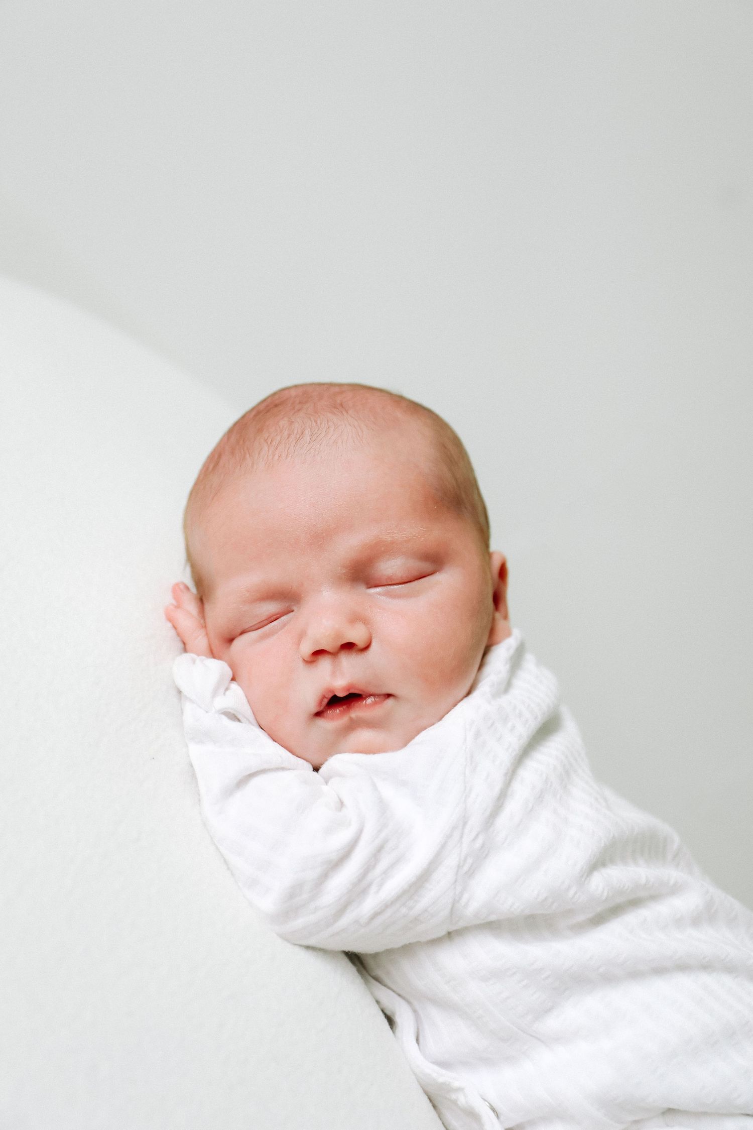 A sleeping newborn baby swaddled in soft white fabric against a white background, during newborn shoot based in Nottingham