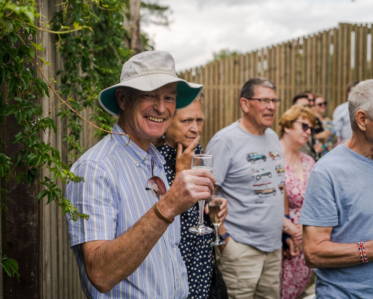 People gather at an outdoor garden or patio space with wooden barrels and greenery in the background.