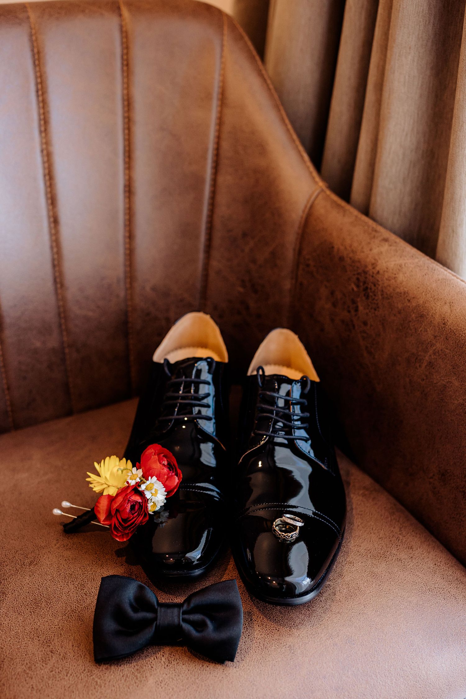 Elegant black patent leather dress shoes and bow tie arranged with red and yellow flowers on brown leather chair.