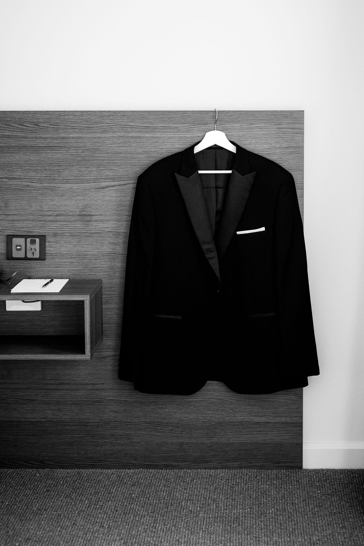 A black suit jacket hangs on a white hanger against a dark wood-paneled wall in a minimalist room.