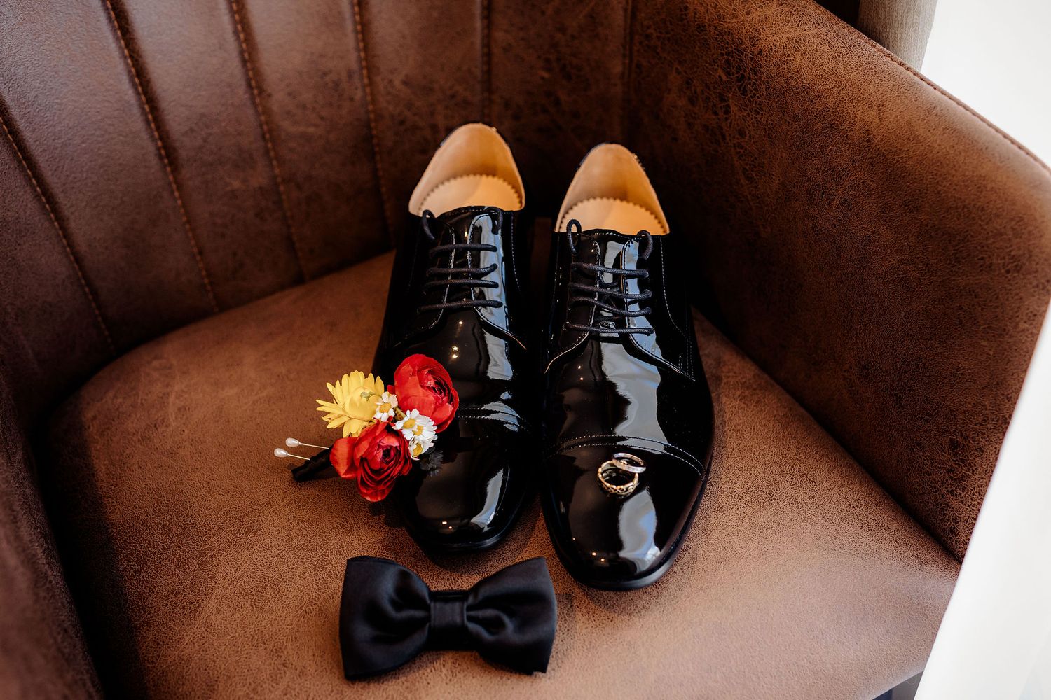 Shiny black formal dress shoes and bow tie placed on brown leather chair with small red and yellow flowers.