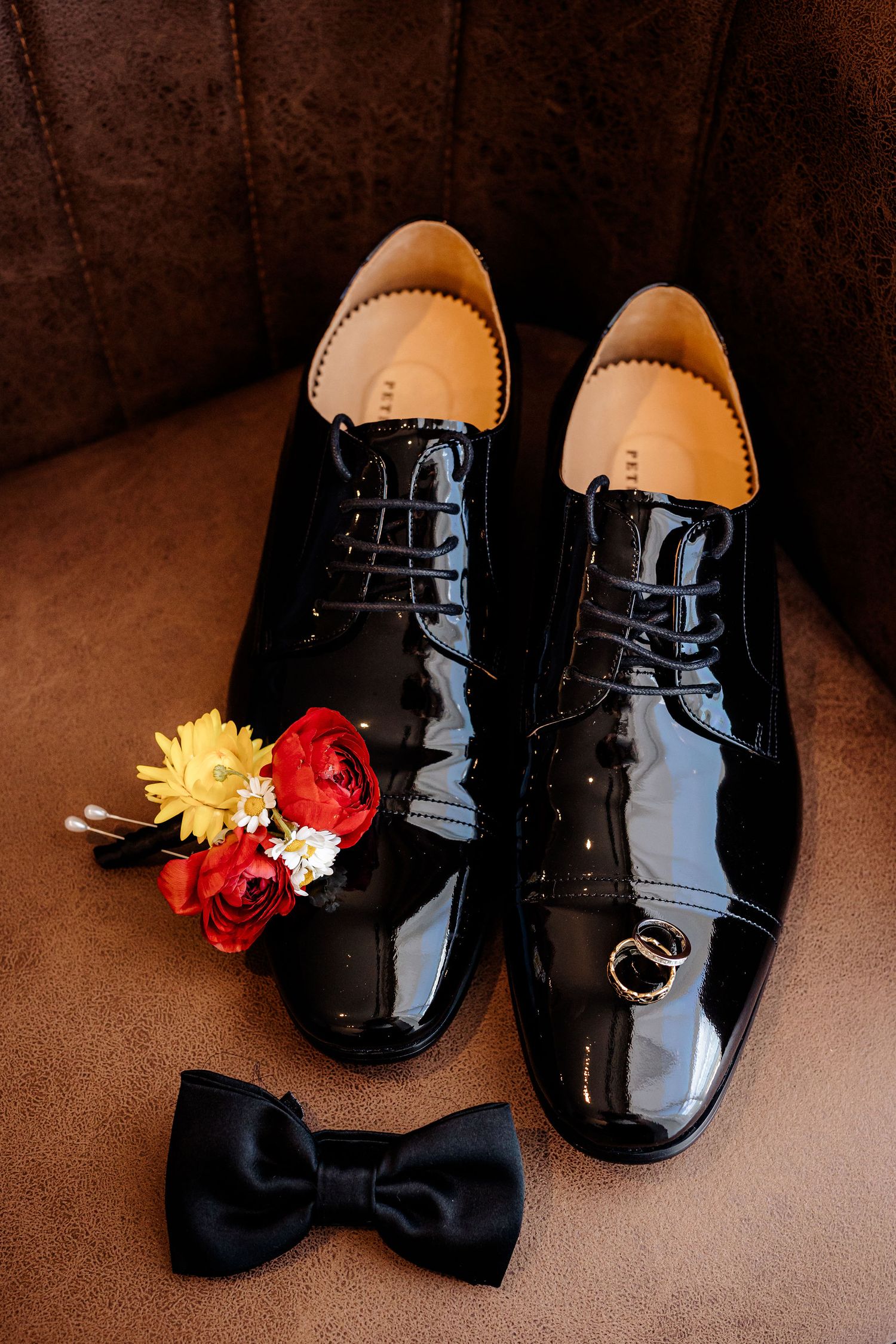 Shiny black patent leather dress shoes with bow tie and red and yellow boutonniere on brown background.