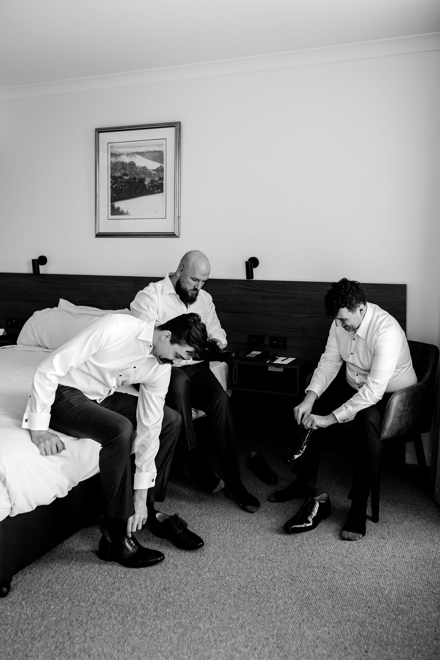Three people in white shirts sit on beds in a hotel room getting ready, with a framed artwork on the wall.