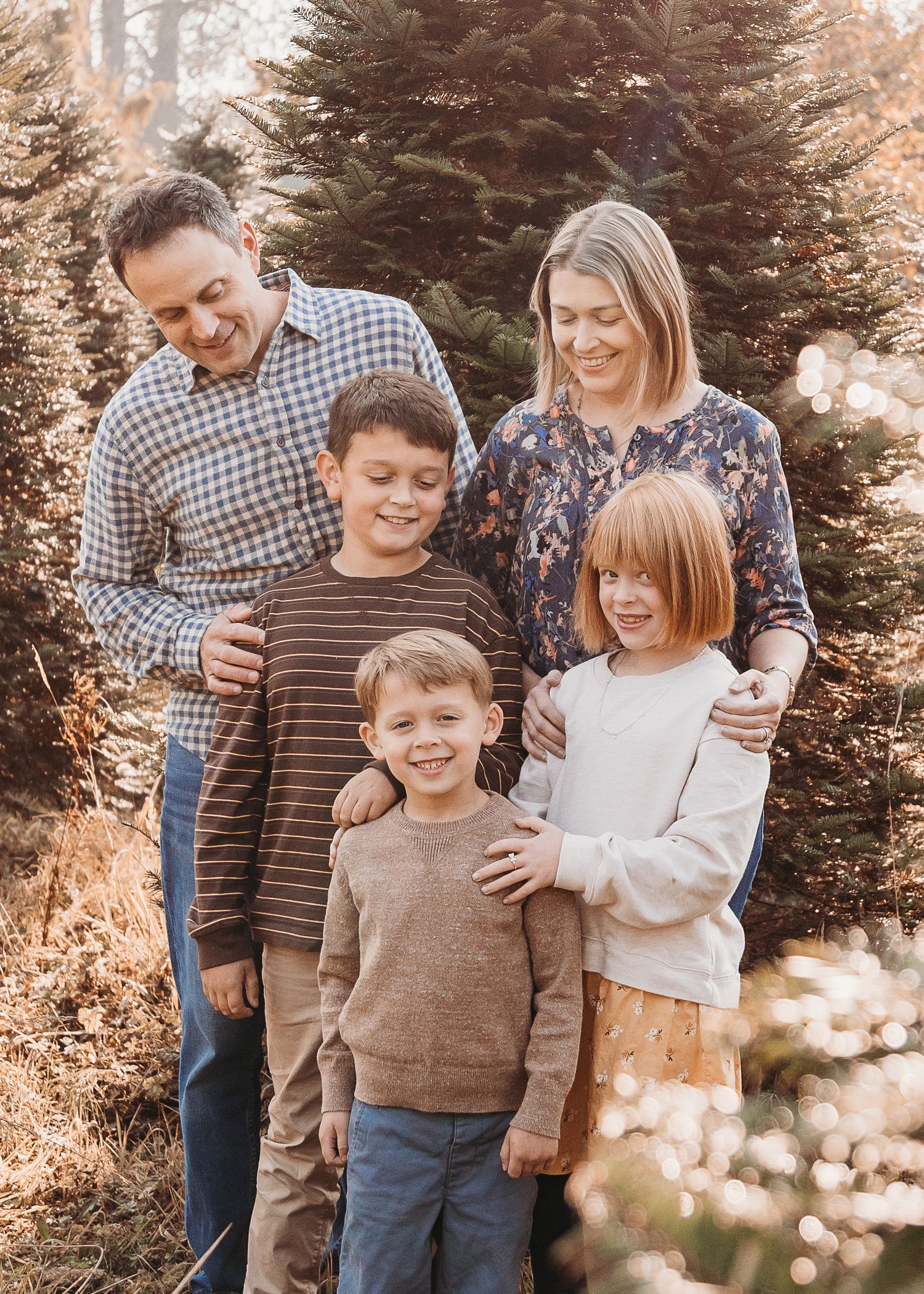 Family poses together outdoors in front of evergreen trees in warm autumn lighting.
