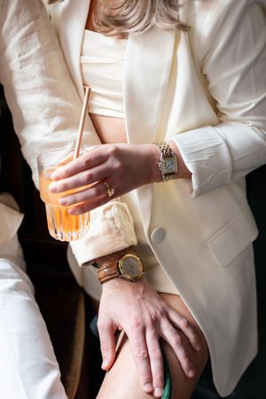 Close-up of hands holding an orange cocktail, showcasing elegant watches and white attire.