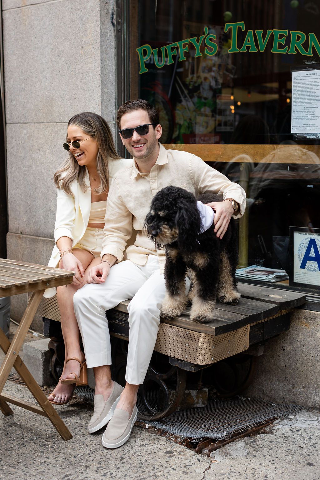 Couple in white outfits sitting with dog outside Puffy's Tavern.