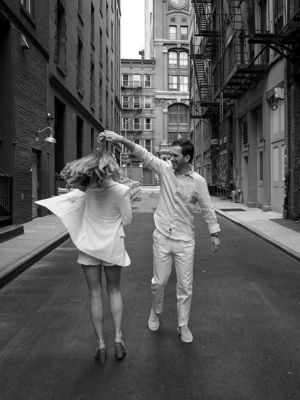 A couple dances playfully in black and white on an urban street with fire escapes and brick buildings.