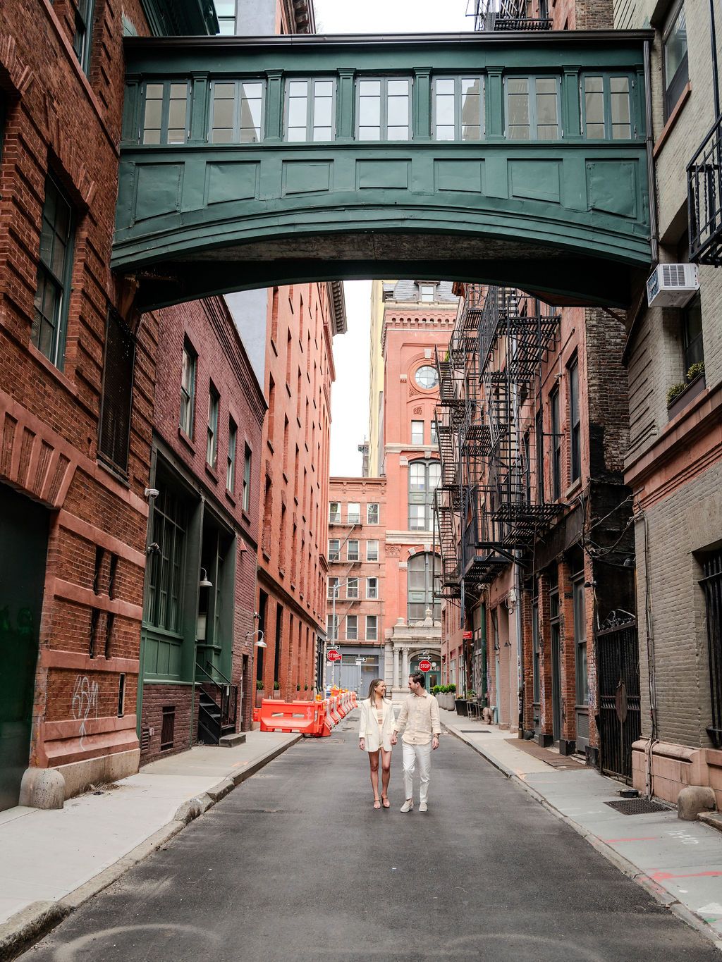 A couple in white outfits walks under a vintage green skybridge connecting historic brick buildings.