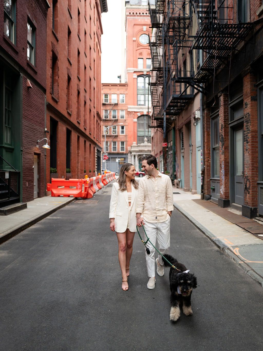 A couple in white outfits stroll down a historic alley with their black dog.