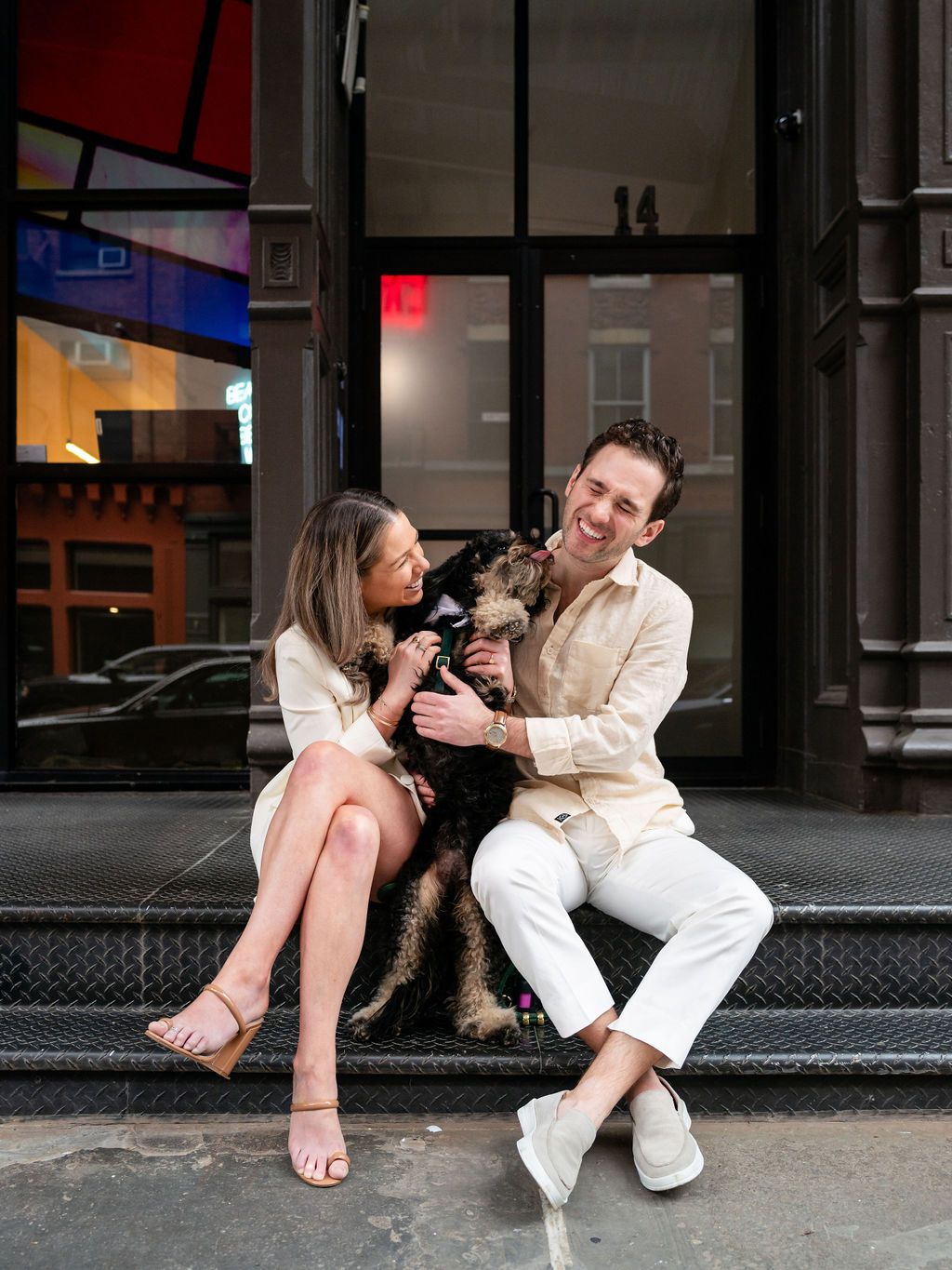 A couple sits on steps with their black dog, sharing a joyful moment together.
