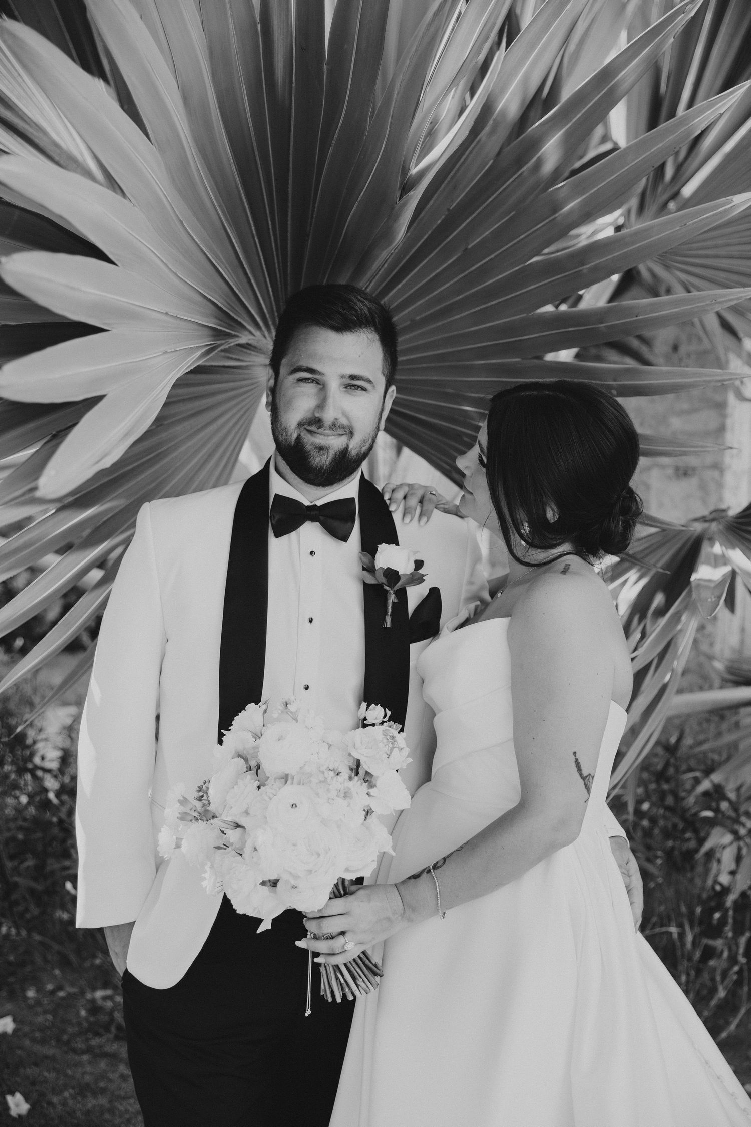 A wedding portrait featuring formal attire and white floral bouquet against palm leaves.