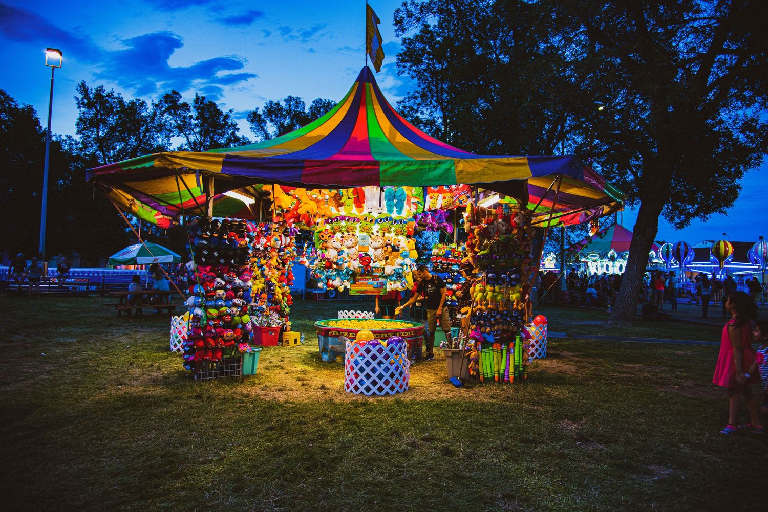 A vibrant carnival game booth with a striped, multi-colored tent and an assortment of plush prizes on display, photographed at dusk.