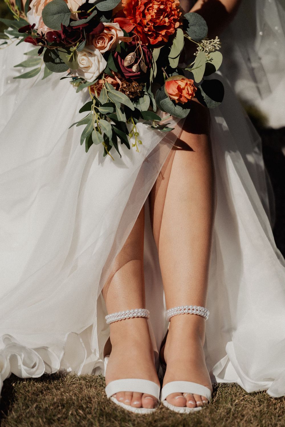 Closeup of white wedding shoes with pearl straps and fall bouquet against flowing dress fabric.